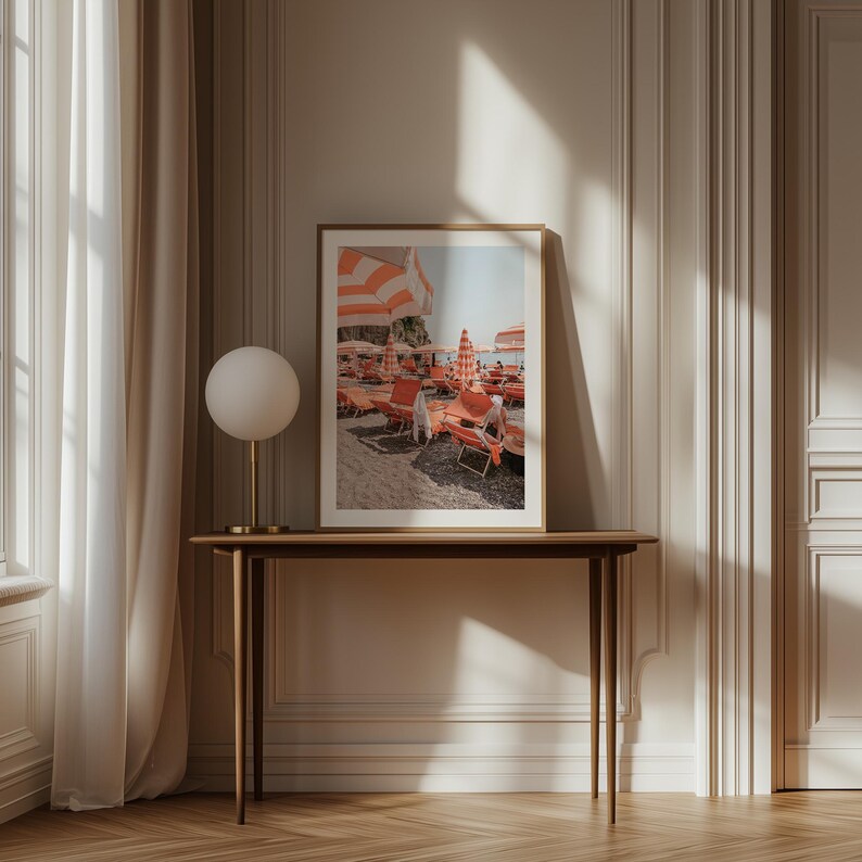 May include: A framed print depicts a beach scene with orange and white striped umbrellas and lounge chairs. The print is displayed on a dark wood table, with a gold-toned lamp and a white globe shade. The room has a neutral color palette.