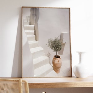 K&ouml;nnte beinhalten: Ein gerahmtes Foto einer wei&szlig;en Treppe mit einem kleinen Olivenbaum in einem Terrakotta-Topf. Die Treppe befindet sich in einem wei&szlig;en Geb&auml;ude mit einer wei&szlig;en Wand im Hintergrund. Das Foto steht auf einem Holztisch mit einer wei&szlig;en Vase und einem Stuhl.