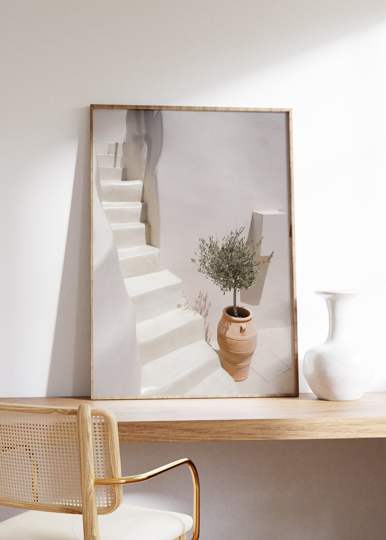 K&ouml;nnte beinhalten: Ein gerahmtes Foto einer wei&szlig;en Treppe mit einem kleinen Olivenbaum in einem Terrakotta-Topf. Die Treppe befindet sich in einem wei&szlig;en Geb&auml;ude mit einer wei&szlig;en Wand im Hintergrund. Das Foto steht auf einem Holztisch mit einer wei&szlig;en Vase und einem Stuhl.