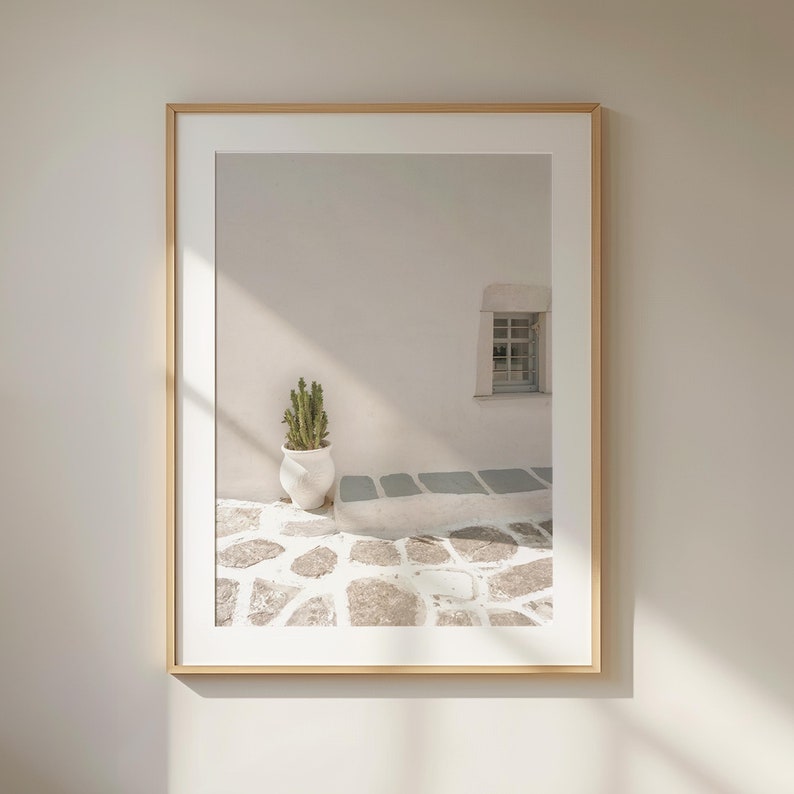 May include: A framed print featuring a minimalist architectural scene. A white wall with a small window and a cactus in a white pot are visible. The ground is paved with irregular stones, and the frame is a light wood color.
