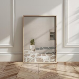 May include: Framed print of a serene outdoor scene. A white wall with a small window and a cactus in a white pot are visible. The ground is paved with light-colored stones, and the frame is a light wood color.