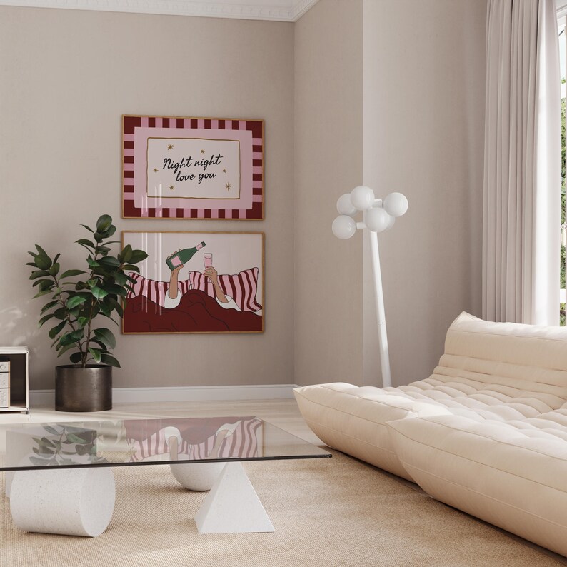 K&ouml;nnte beinhalten: Zwei gerahmte Kunstdrucke h&auml;ngen an einer Wand. Der obere Druck hat den Text "Night night love you" in einem rosa und bordeauxfarbenen Rahmen. Der untere Druck zeigt eine Person, die Champagner einschenkt. Ein modernes Wohnzimmer mit einem Glastisch und einem beigen Sofa.