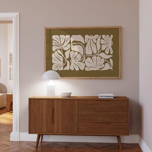 May include: A framed floral art print with a light wood frame hangs above a mid-century modern wooden cabinet. The artwork features white and pink floral shapes on an olive green background. A white lamp and bowl sit on the cabinet.