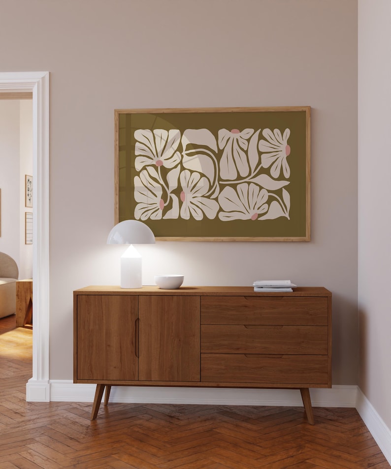 May include: A framed floral art print with a light wood frame hangs above a mid-century modern wooden cabinet. The artwork features white and pink floral shapes on an olive green background. A white lamp and bowl sit on the cabinet.