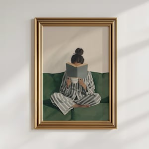 May include: Framed art print of a person in striped pajamas reading a book while seated on a green sofa. The person is holding a white cup. The artwork is in a gold frame and is against a white wall.
