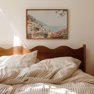 May include: A framed print of a coastal Italian town with colorful buildings, set against a blue sea and sky. The print is mounted above a bed with striped bedding and a wooden headboard.