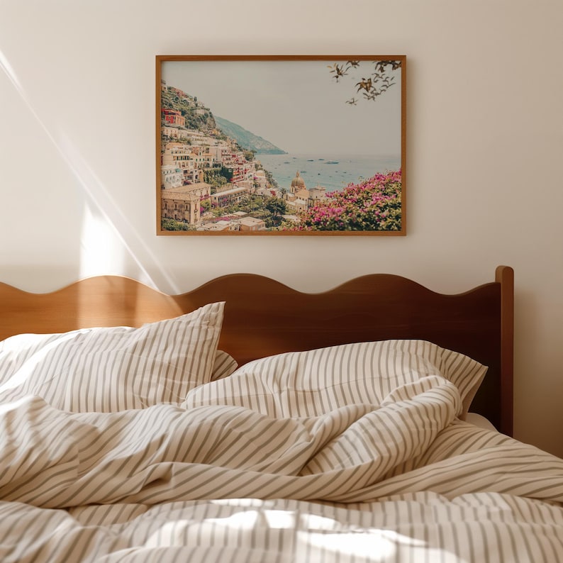 May include: A framed print of a coastal Italian town with colorful buildings, set against a blue sea and sky. The print is mounted above a bed with striped bedding and a wooden headboard.