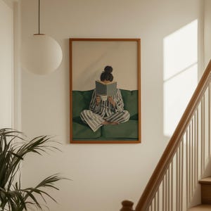May include: A framed print depicts a person in striped pajamas reading a book while seated on a green sofa. The person holds a cup, suggesting a moment of relaxation. The artwork is displayed on a white wall, with a round light fixture and a staircase visible.