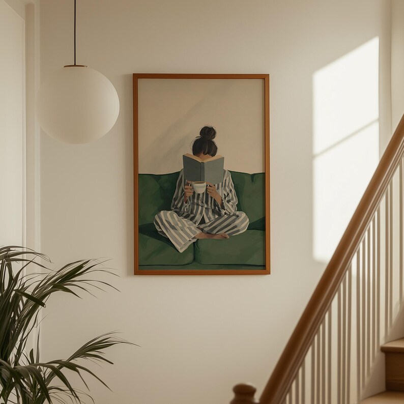 May include: A framed print depicts a person in striped pajamas reading a book while seated on a green sofa. The person holds a cup, suggesting a moment of relaxation. The artwork is displayed on a white wall, with a round light fixture and a staircase visible.