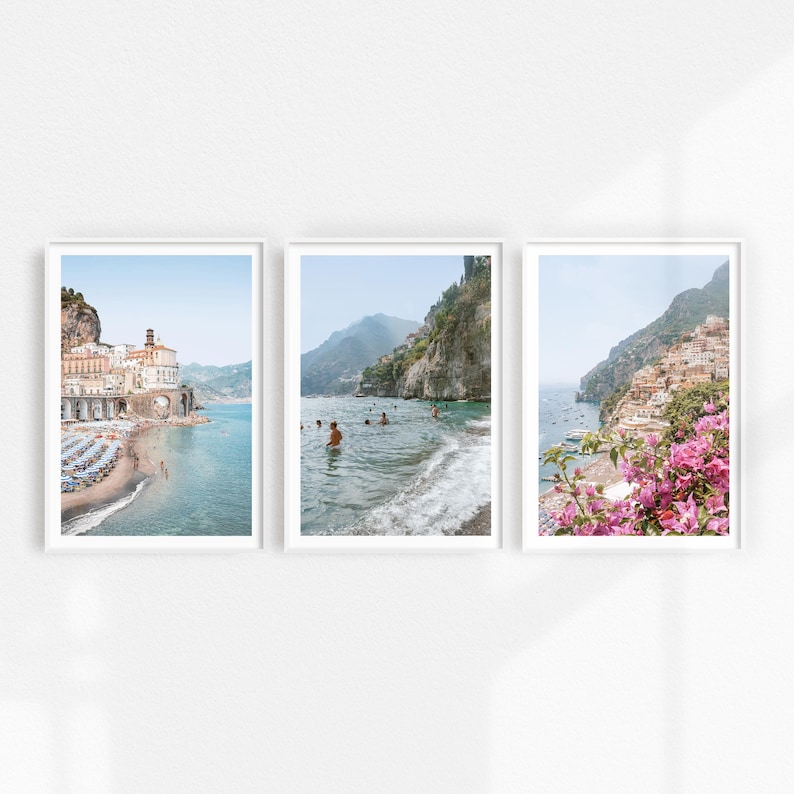 May include: Three framed prints of coastal scenes. The first print shows a white building with a blue roof on a rocky cliff overlooking a turquoise ocean. The second print shows a rocky cliff with a turquoise ocean and people swimming. The third print shows a rocky cliff with a turquoise ocean and pink flowers in the foreground.