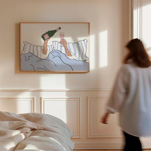 May include: A framed illustration of a person in bed with a glass of champagne and a bottle of champagne. The person is holding the glass in one hand and the bottle in the other. The illustration is in a minimalist style with a light blue background.