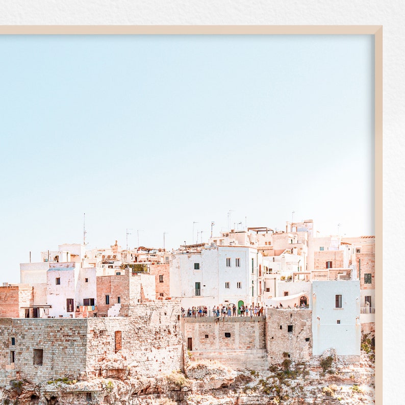 May include: A whitewashed village perched on a cliff overlooking a blue sky. The buildings are clustered together, with a few trees and shrubs growing on the cliffside. A group of people are standing on a ledge overlooking the sea.