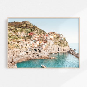 May include: A colorful Italian village built into a cliffside overlooking a turquoise bay. The village is a mix of pastel yellow, pink, orange, and red buildings with terracotta roofs. The water is clear and calm, with a small boat in the foreground.