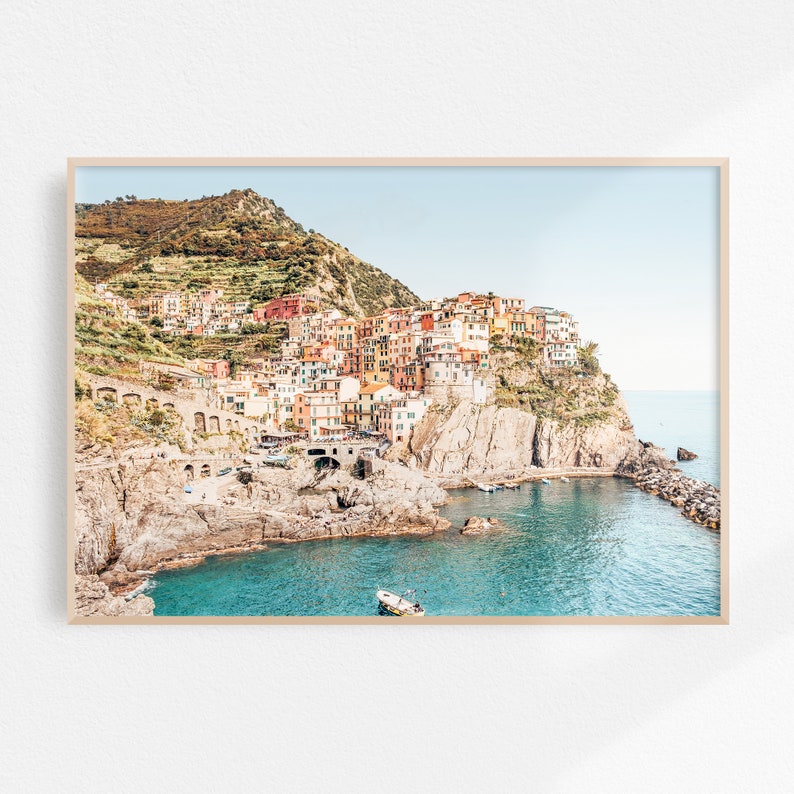 May include: A colorful Italian village built into a cliffside overlooking a turquoise bay. The village is a mix of pastel yellow, pink, orange, and red buildings with terracotta roofs. The water is clear and calm, with a small boat in the foreground.