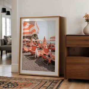 May include: Framed print of a beach scene with orange and white striped umbrellas and lounge chairs. The photograph captures a sunny day at the beach, with a view of the ocean and a rocky cliffside. The frame is a light wood color.