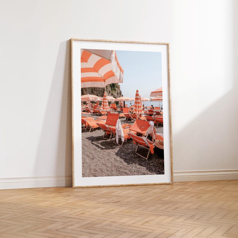 May include: A framed print of a beach scene with orange and white striped umbrellas and lounge chairs. The beach is filled with people enjoying the sun. The print is displayed on a wooden floor.
