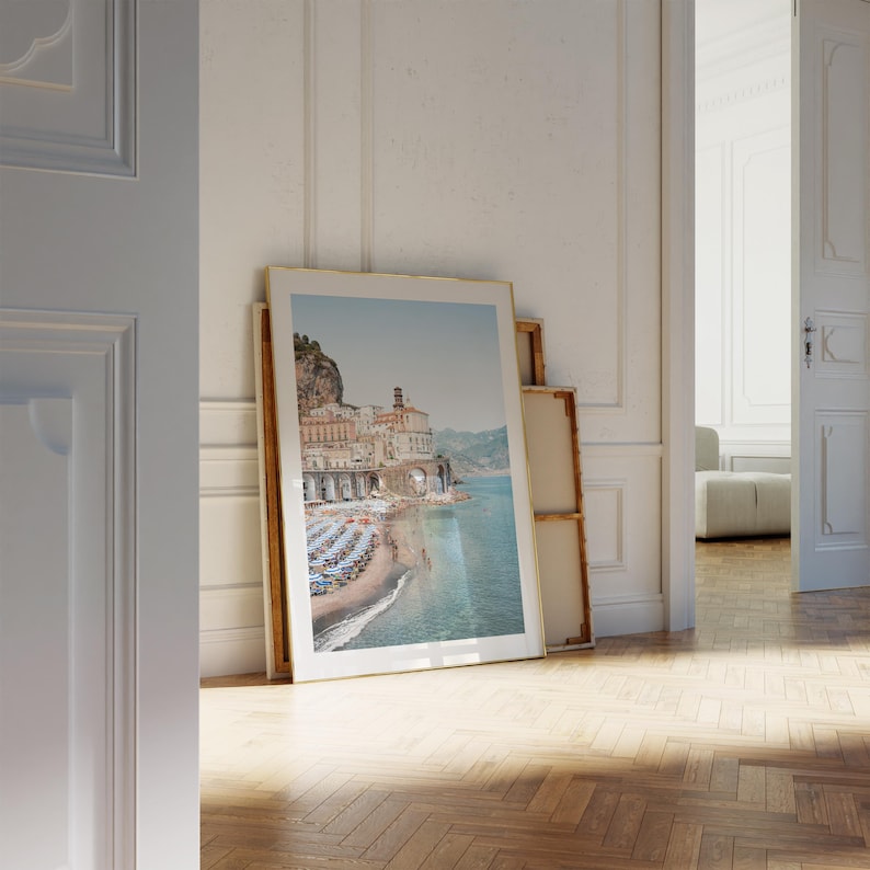 K&ouml;nnte beinhalten: Ein gerahmtes Foto einer K&uuml;stenstadt mit bunten Geb&auml;uden, einem Sandstrand und blauen Sonnenschirmen. Das Foto h&auml;ngt an einer wei&szlig;en Wand in einem Raum mit Holzboden.