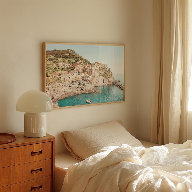 May include: A framed photograph of a colorful town built on a cliffside overlooking a turquoise ocean. The photo is hanging on a white wall above a bed with white linens.