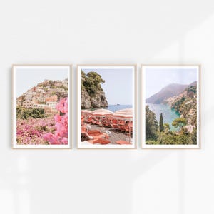 Pu&ograve; includere: Tre stampe incorniciate di paesaggi costieri italiani. La prima stampa mostra una citt&agrave; in collina con fiori rosa in primo piano. La seconda stampa mostra una spiaggia con ombrelloni e sedie a strisce rosse e bianche. La terza stampa mostra una costa rocciosa con un mare turchese.