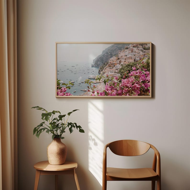 May include: A framed photograph of a coastal town with colorful buildings and a view of the ocean. Pink flowers are in the foreground.