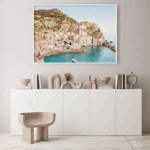 May include: A framed photograph of a colorful village built into a cliffside overlooking a turquoise ocean. The village is surrounded by lush green hills and the sky is a light blue.