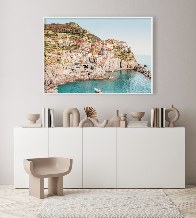 May include: A framed photograph of a colorful village built into a cliffside overlooking a turquoise ocean. The village is surrounded by lush green hills and the sky is a light blue.