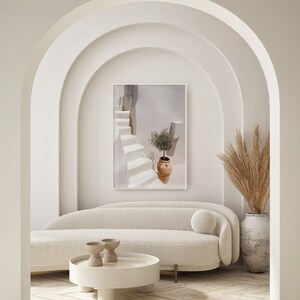 K&ouml;nnte beinhalten: Ein wei&szlig;es Sofa mit einem wei&szlig;en Kissen steht in einem Raum mit wei&szlig;en W&auml;nden und einem Bogenportal. Ein wei&szlig;er Couchtisch mit runder Platte steht vor dem Sofa. Zwei kleine braune Keramikschalen stehen auf dem Tisch. Ein gerahmtes Foto einer wei&szlig;en Treppe mit einer Topfpflanze h&auml;ngt an der Wand &uuml;ber dem Sofa. Eine hohe Vase mit getrocknetem Pampasgras steht auf dem Boden neben dem Sofa.