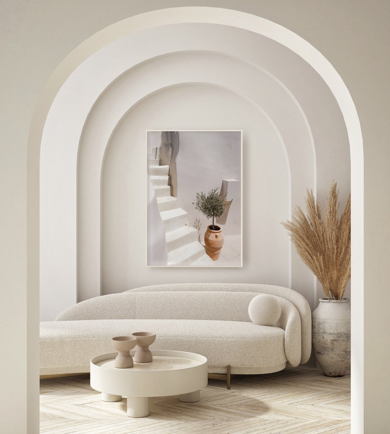 K&ouml;nnte beinhalten: Ein wei&szlig;es Sofa mit einem wei&szlig;en Kissen steht in einem Raum mit wei&szlig;en W&auml;nden und einem Bogenportal. Ein wei&szlig;er Couchtisch mit runder Platte steht vor dem Sofa. Zwei kleine braune Keramikschalen stehen auf dem Tisch. Ein gerahmtes Foto einer wei&szlig;en Treppe mit einer Topfpflanze h&auml;ngt an der Wand &uuml;ber dem Sofa. Eine hohe Vase mit getrocknetem Pampasgras steht auf dem Boden neben dem Sofa.