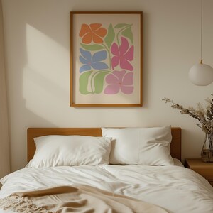 K&ouml;nnte beinhalten: Ein gerahmter Kunstdruck mit orangefarbenen, blauen und rosa Blumen und gr&uuml;nen Bl&auml;ttern. Das Kunstwerk h&auml;ngt &uuml;ber einem Bett mit wei&szlig;er Bettw&auml;sche und einem h&ouml;lzernen Kopfteil und schafft eine ruhige Schlafzimmer&auml;sthetik.