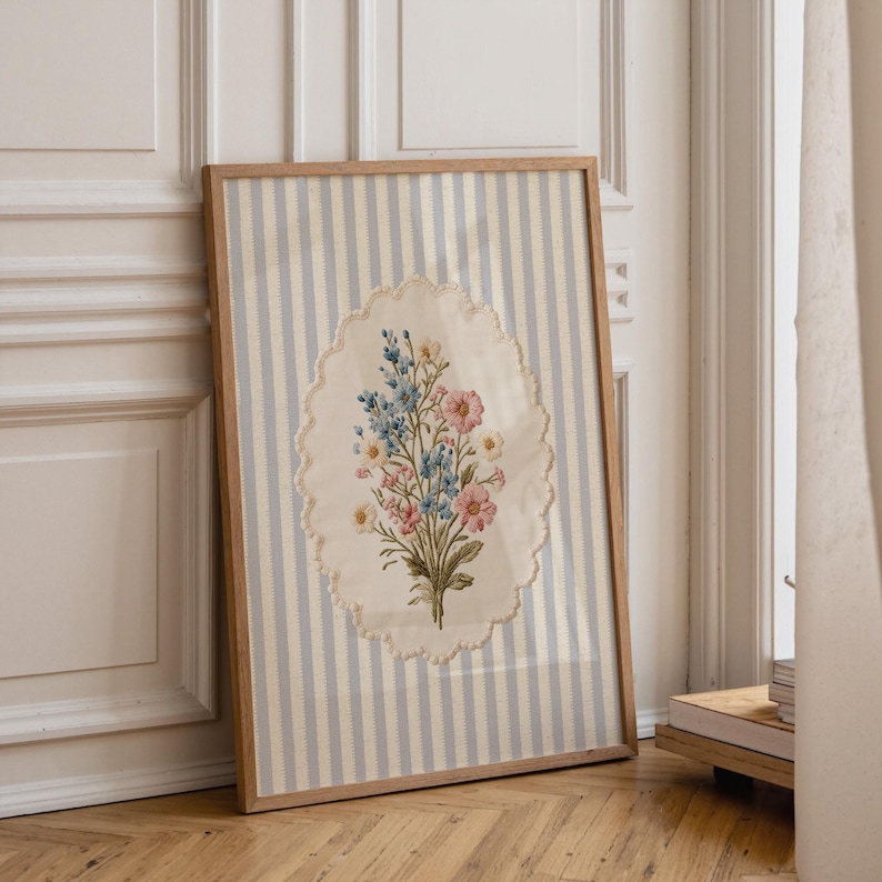 May include: A framed floral embroidery. The artwork features a bouquet of pink, blue, and yellow flowers on an oval-shaped, cream-colored background with a scalloped edge. The background is a light blue and white striped fabric, encased in a wooden frame.