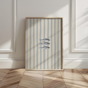 May include: A framed art print featuring three stylized fish illustrations. The artwork is set against a background of vertical blue and cream stripes. The frame is a light wood color, and the overall aesthetic is coastal and minimalist.