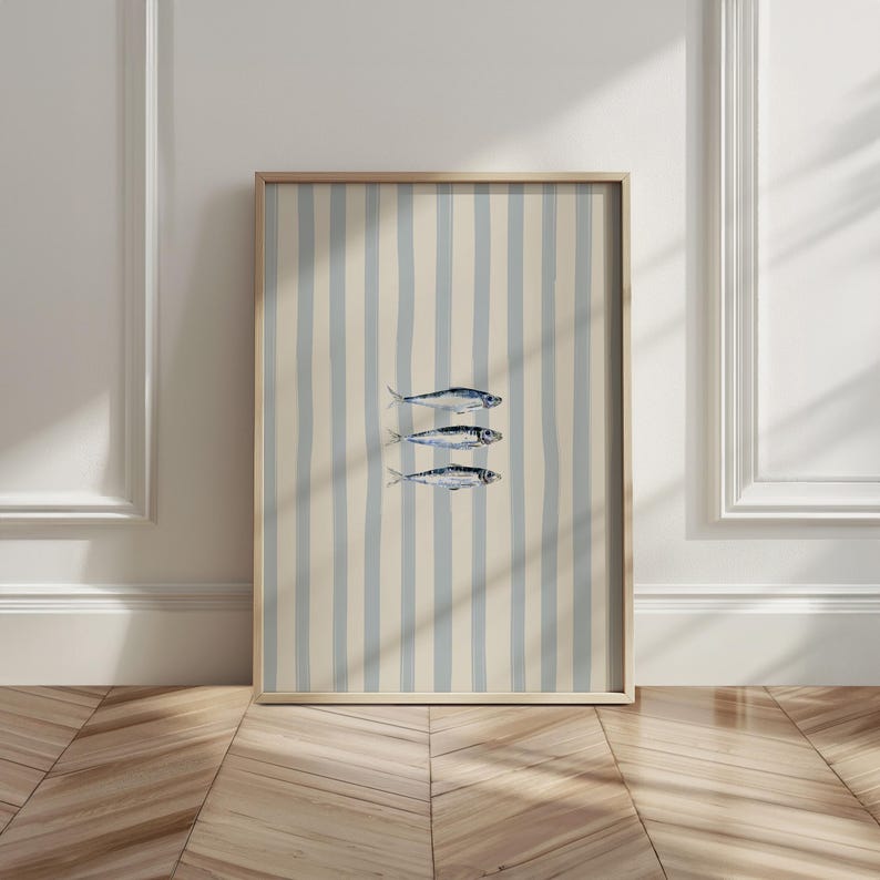 May include: A framed art print featuring three stylized fish illustrations. The artwork is set against a background of vertical blue and cream stripes. The frame is a light wood color, and the overall aesthetic is coastal and minimalist.