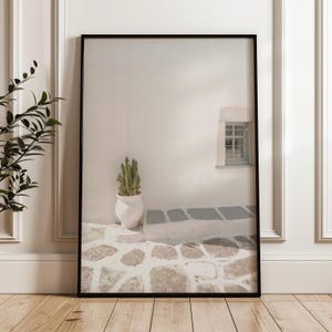 May include: Framed print featuring a minimalist architectural scene. A white wall with a small window and a cactus in a white pot are visible. The ground is paved with stones. The print is in a black frame and leans against a white wall.