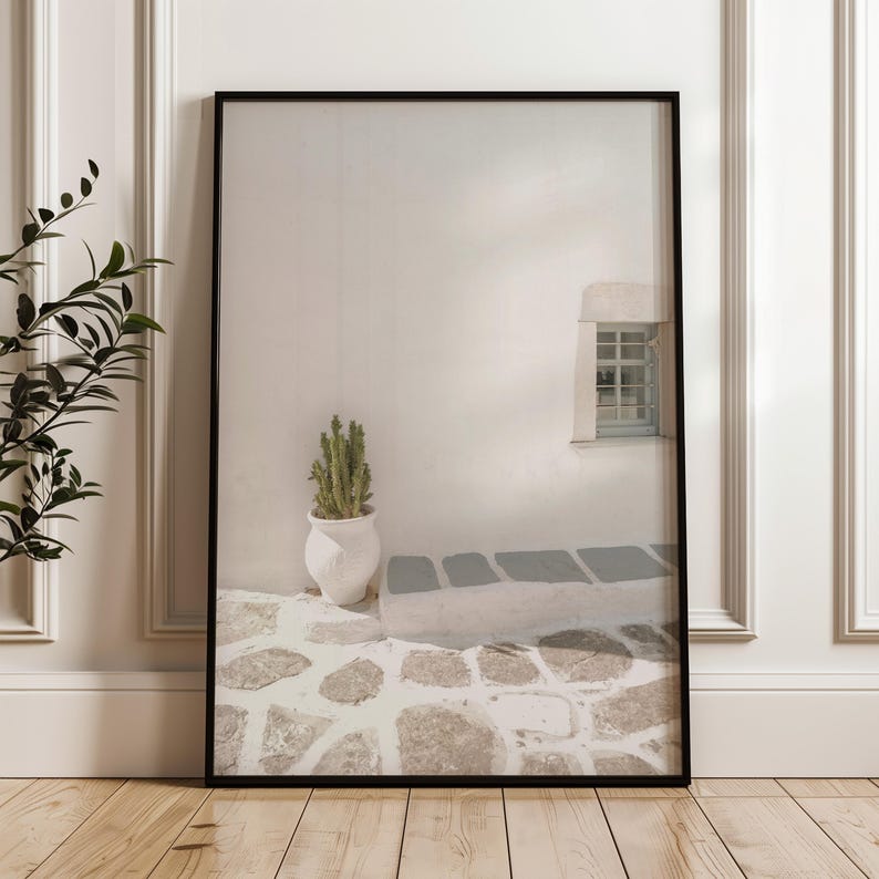 May include: Framed print featuring a minimalist architectural scene. A white wall with a small window and a cactus in a white pot are visible. The ground is paved with stones. The print is in a black frame and leans against a white wall.
