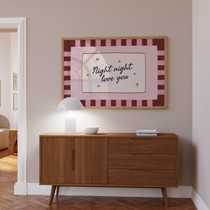 May include: A framed print with the words "Night night love you" in a cursive font, surrounded by stars. The print has a pink and burgundy color scheme and is displayed above a wooden cabinet with a white lamp and a small bowl.