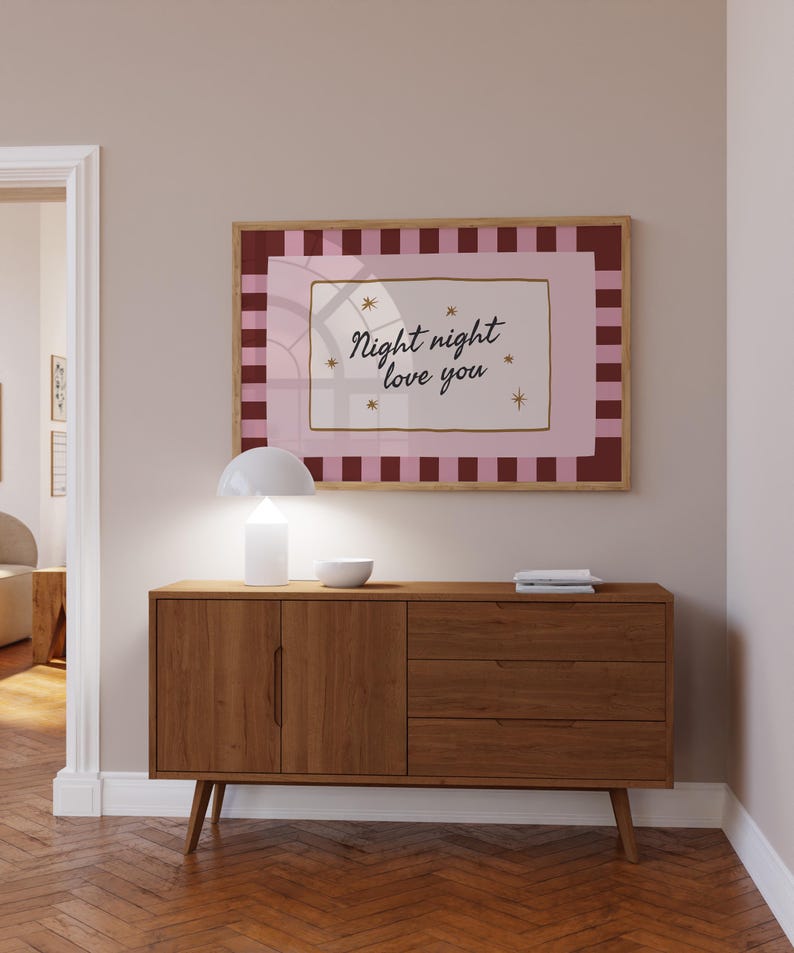 May include: A framed print with the words "Night night love you" in a cursive font, surrounded by stars. The print has a pink and burgundy color scheme and is displayed above a wooden cabinet with a white lamp and a small bowl.