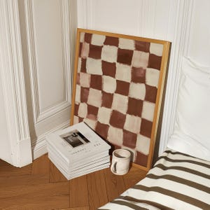 May include: A framed abstract painting with a checkered pattern in shades of brown and white. The painting is leaning against a stack of white magazines with the text "ARK JOURNAL" on the cover. A ceramic mug is sitting on a wooden floor.