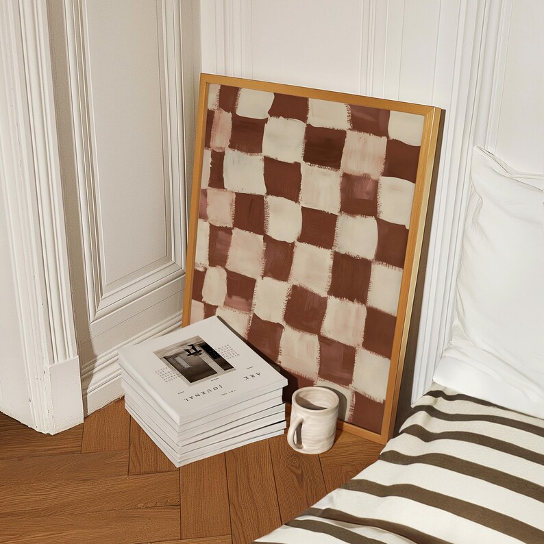 May include: A framed abstract painting with a checkered pattern in shades of brown and white. The painting is leaning against a stack of white magazines with the text "ARK JOURNAL" on the cover. A ceramic mug is sitting on a wooden floor.
