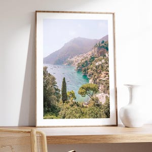 May include: A framed photograph of a coastal landscape with a mountain range in the background, a turquoise blue ocean, and lush green trees in the foreground.