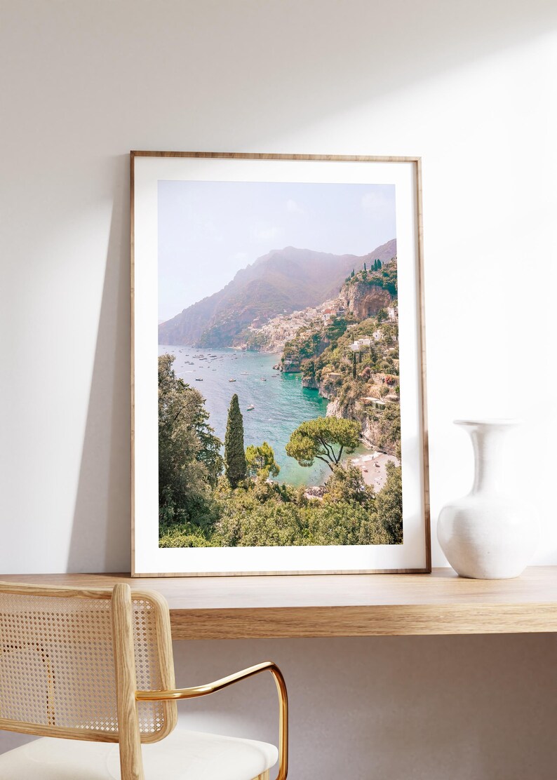 May include: A framed photograph of a coastal landscape with a mountain range in the background, a turquoise blue ocean, and lush green trees in the foreground.