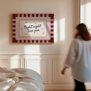 May include: A framed wall art print with the text "Night night love you" in a cursive font. The artwork features a pink and burgundy striped border, with a gold frame. The print is hanging on a white wall in a bedroom.