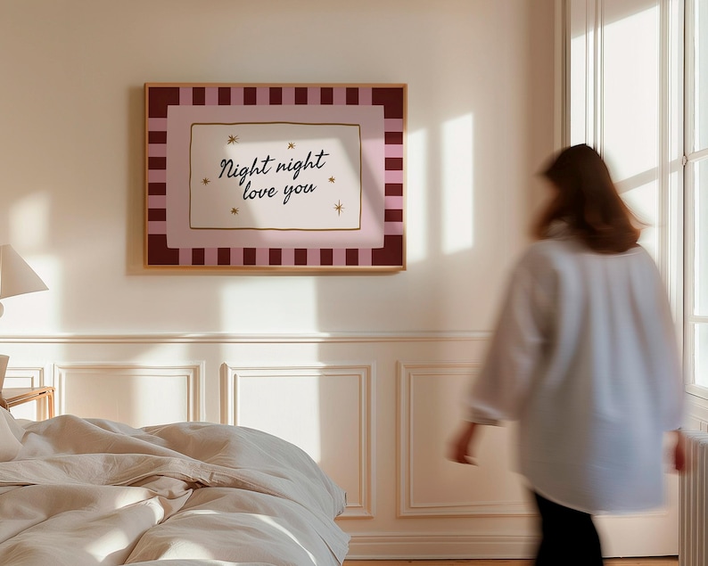 May include: A framed wall art print with the text "Night night love you" in a cursive font. The artwork features a pink and burgundy striped border, with a gold frame. The print is hanging on a white wall in a bedroom.