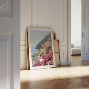 K&ouml;nnte beinhalten: Ein gerahmtes Foto einer K&uuml;stenstadt mit bunten H&auml;usern, die in einen H&uuml;gel gebaut sind. Die Stadt ist von &uuml;ppiger gr&uuml;ner Vegetation und einem blauen Meer umgeben. Im Vordergrund befinden sich rosa Blumen.