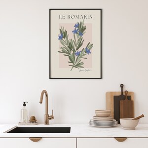 Op de afbeelding: Een zwart ingelijste print met een botanische illustratie van rozemarijn met blauwe bloemen op een lichtroze achtergrond. De titel "LE ROMARIN" staat bovenaan de print.