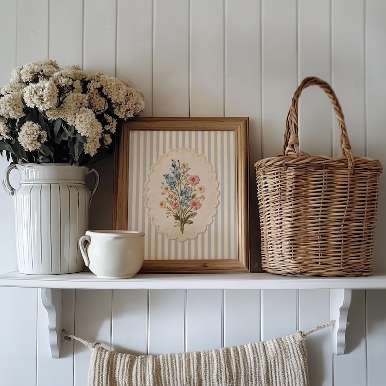 K&ouml;nnte beinhalten: Ein dekoratives Regal zeigt einen gerahmten Blumendruck, einen Weidenkorb, eine wei&szlig;e Keramikvase mit Blumen und eine kleine Keramiktasse. Der Druck hat einen hellblau-wei&szlig; gestreiften Hintergrund.