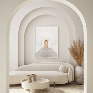 May include: A white couch with a white throw pillow sits in front of a framed print of a white archway with a wooden door. A white coffee table with two small brown ceramic bowls sits in front of the couch. A tall vase with dried beige pampas grass sits on the right side of the image.