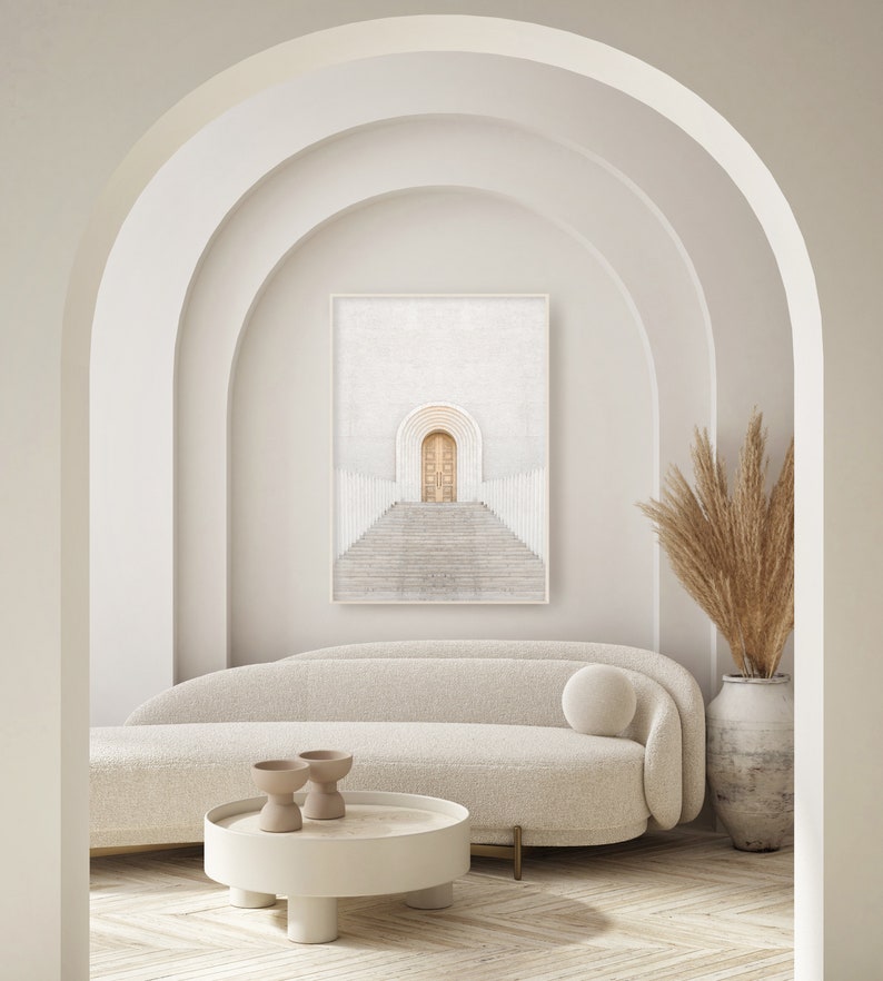 May include: A white couch with a white throw pillow sits in front of a framed print of a white archway with a wooden door. A white coffee table with two small brown ceramic bowls sits in front of the couch. A tall vase with dried beige pampas grass sits on the right side of the image.