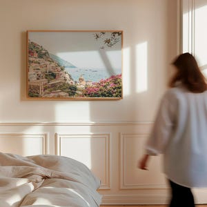 May include: A framed print of a coastal landscape with a town built into a hillside. The print is hanging on a white wall in a room with a bed and a window.