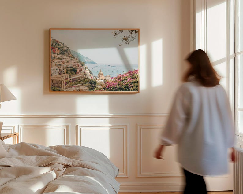 May include: A framed print of a coastal landscape with a town built into a hillside. The print is hanging on a white wall in a room with a bed and a window.