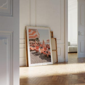 May include: A framed print of a beach scene with orange and white striped umbrellas and lounge chairs. The print is leaning against a wall in a room with a wooden floor. The image evokes a sunny, relaxing atmosphere.